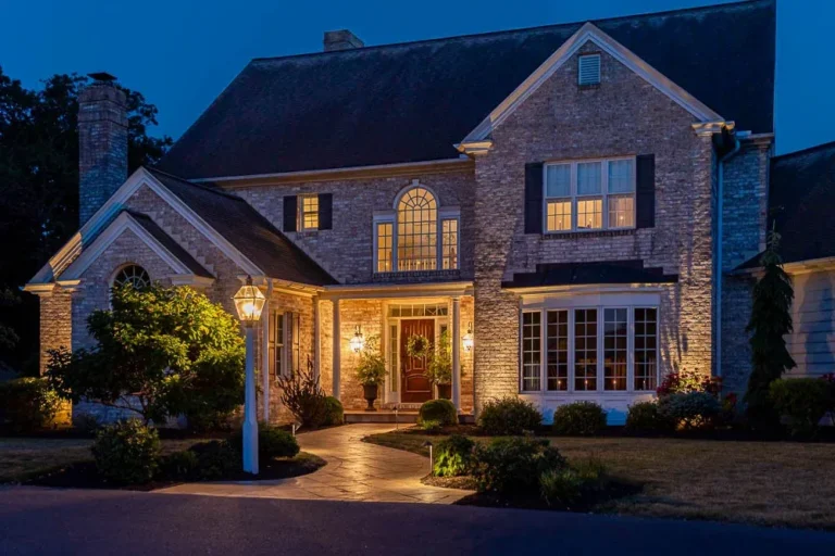 Elegant home exterior at night featuring well-lit entryway, landscaped gardens, and inviting pathway, showcasing outdoor lighting and hardscape design by Authentic Homescapes.