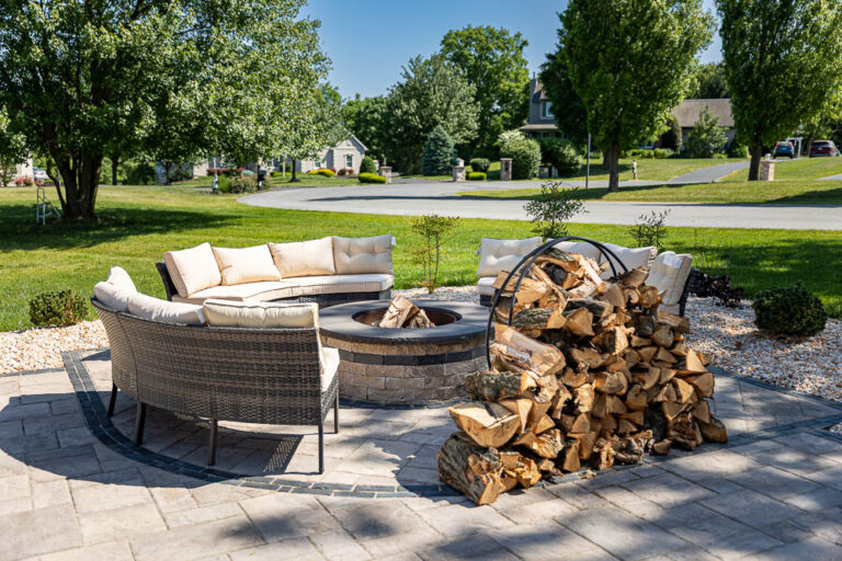 Outdoor living space featuring a circular seating area with plush cushions around a stone fire pit, stacked firewood nearby, surrounded by greenery and trees, ideal for relaxation and hosting gatherings.
