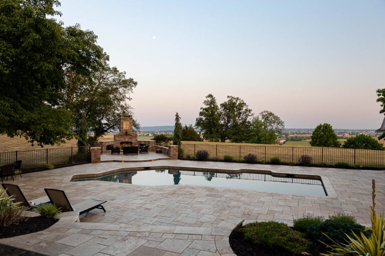 Elegant outdoor pool area with stone patio, lounge chairs, and fireplace, surrounded by lush greenery, showcasing a functional and beautiful landscape design by Authentic Homescapes in Denver, PA.