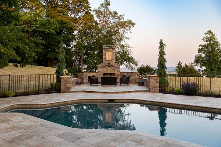 Outdoor living space featuring a stone fireplace and seating area beside a pool, surrounded by greenery, showcasing high-quality hardscaping and landscaping solutions by Authentic Homescapes in Denver, PA.