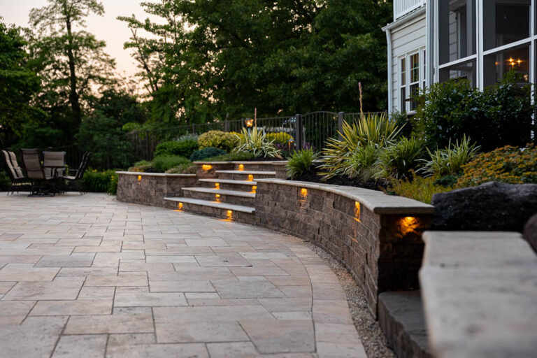 Curved stone retaining wall with integrated lighting, leading to a landscaped outdoor space featuring paver patio, steps, and lush greenery, enhancing hardscaping design for a beautiful backyard.