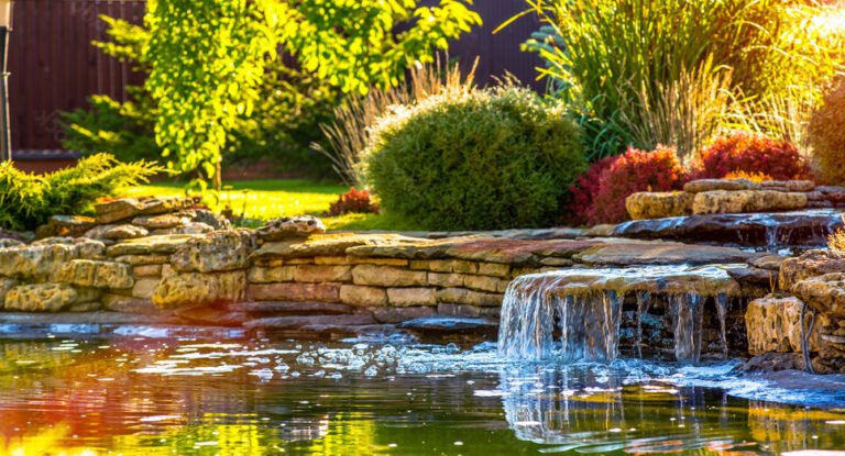 Water feature with cascading waterfall and stone border, surrounded by lush greenery and colorful plants, enhancing outdoor tranquility and relaxation.