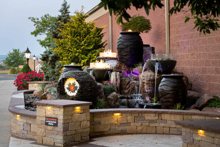 Outdoor water feature with stacked stone walls, decorative urns, and illuminated plants, enhancing a landscaped area for functional outdoor spaces in Denver, PA.