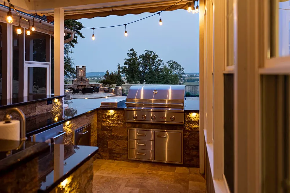 Covered outdoor kitchen beside a pool at dusk with string and counter lights on